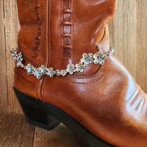 May include: A silver flower boot cuff with a chain link design. The cuff is worn on a brown leather cowboy boot.