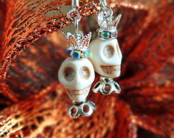 May include: A pair of skull earrings with silver-tone metal accents. Each skull is cream-colored and topped with a silver crown adorned with iridescent rhinestones. The earrings have a decorative base and are suspended from silver hooks.