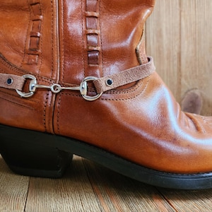 May include: Brown leather cowboy boot with a brown leather strap and silver buckle. The strap is attached to the boot with two small silver rings.