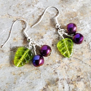 May include: Dangle earrings with a cherry design. Each earring has three iridescent purple beads and a green leaf-shaped bead. The earrings are suspended from silver-coloured fish hook ear wires.