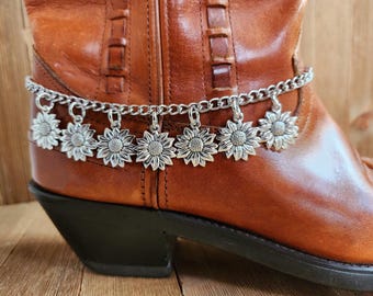 May include: A silver-colored boot chain with sunflower charms is draped over a brown leather boot. The chain is made of small, linked metal pieces, and the sunflower charms are detailed with petals and a central disc.