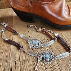 May include: Two silver and turquoise necklaces with feather accents and arrow charms. The necklaces are on a wooden surface.