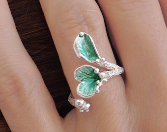 Puede incluir: Un anillo de plata con dos elementos abiertos en forma de hoja con detalles de esmalte verde vibrante. La banda tiene una apariencia texturizada similar a una rama con pequeñas cuentas de plata en los extremos, creando un diseño abierto.