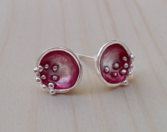 May include: A pair of silver stud earrings with a unique design. Each earring features a round, open-faced shape with a vibrant magenta interior and small, silver bead accents. The earrings are set against a light wooden surface.