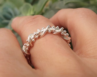 May include: A silver ring with a twisted vine design, adorned with small, round beads. The ring is held by a hand, with a blurred green background of foliage.
