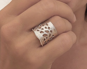 May include: A wide silver-toned ring featuring an intricate, organic cut-out pattern with a textured, brushed metal finish. The band has a slightly irregular, wavy edge design that wraps around the finger, showcasing a detailed, perforated surface.