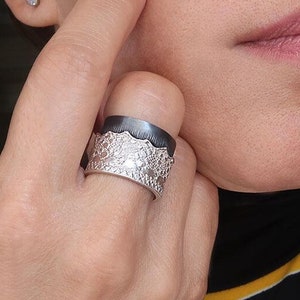 Op de afbeelding: Een zilveren ring met een getextureerd, zwart en zilver ontwerp. De ring is breed en heeft een unieke, organische vorm.