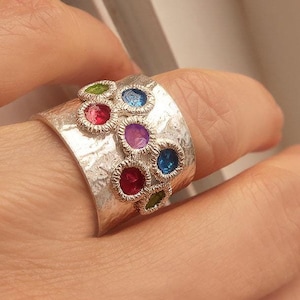 May include: A silver ring with a hammered texture and colorful gemstones. The ring features a cluster of round, multi-colored stones set in silver.