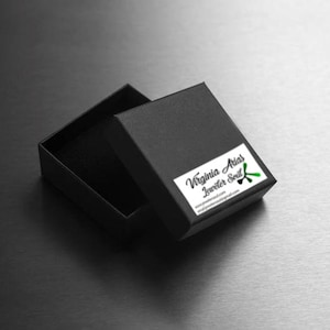 May include: A black jewelry box with a green and black logo that says "Virginia Arias Jeweler Soul" and the website address "virginiaariasjewelry.com".