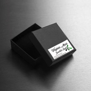 May include: An open black jewelry box with a white label featuring the text "Virginia Arias Jeweler Soul" and a green accent. The box is on a dark surface.