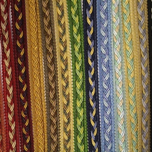 May include: A close-up image of various braided trim ribbons in a spectrum of colours. The ribbons are arranged vertically, displaying a range of hues from red and gold to blue and black. Each ribbon has a unique braided pattern.