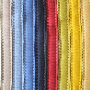 May include: A close-up of a selection of colourful fringe trim in various shades. The colours include white, beige, grey, blue, navy, red, yellow, and black. Each strip has a stitched edge and a fringed bottom.