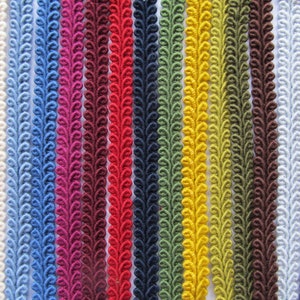 May include: A selection of decorative trim in a spectrum of colours, such as white, blue, red, and green. Each trim displays a repeating, leaf-like design. The trims are arranged vertically, demonstrating the variety of shades suitable for crafting and design applications.