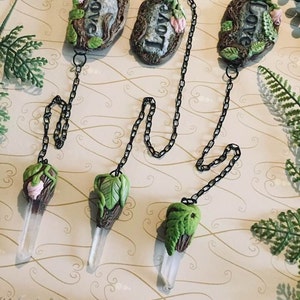 May include: Three crystal point pendants with a green and brown clay design, each with a small pink flower. The pendants are attached to black chains with two grey clay charms that say "Love" in white lettering.