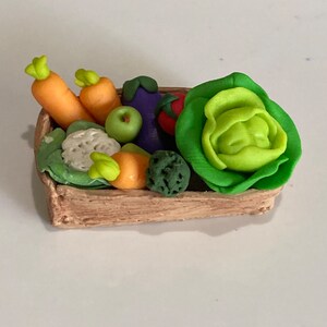 May include: A small wooden crate filled with a variety of colorful vegetables and fruit, including carrots, an apple, a cauliflower, a green pepper, an eggplant, a tomato, and a head of lettuce.