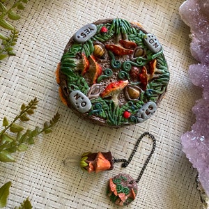 May include: A circular divination board made of wood, featuring a forest scene with mushrooms, leaves, and stones. The board is divided into four sections labelled "Yes", "No", "Maybe", and "On". A pendulum with a mushroom charm hangs from a black chain.