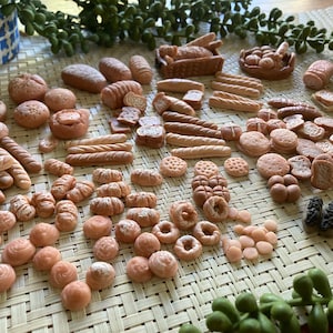 May include: A variety of miniature baked goods, including loaves of bread, rolls, croissants, and pastries, are arranged on a woven mat. The miniature baked goods are made of polymer clay and are painted in shades of brown and beige.