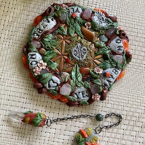 May include: A circular clay board with a green, brown, and orange design. The board is decorated with various stones and crystals, including rose quartz, amethyst, and clear quartz. The board also features the words "Believe", "Love", "Yes", and "Always". A clear quartz pendulum with a brown and orange clay top is hanging from a black chain.