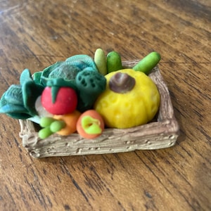 May include: A small, brown, woven basket filled with miniature vegetables. The basket contains a yellow squash, a red tomato, a green leafy vegetable, and a few small carrots.