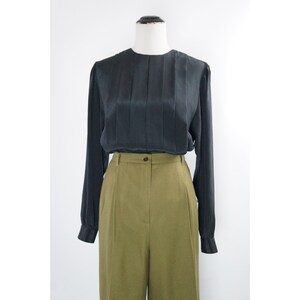 Könnte beinhalten: Eine schwarze Bluse mit langen Ärmeln, einer gefalteten Vorderseite und einem runden Ausschnitt. Die Bluse wird mit einer olivgrünen, hochgeschnittenen Hose kombiniert.