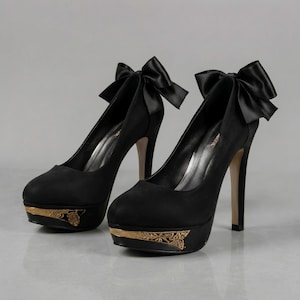 May include: A pair of black high-heeled pumps featuring a rounded toe, a platform sole with gold-toned floral filigree detailing, and a large black satin bow attached to the back of the heel. The interior lining is silver with visible branding.