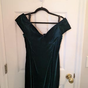 May include: A dark green velvet dress with an off-the-shoulder neckline hangs on a black hanger. The dress has a fitted bodice and a long, flowing skirt. The fabric appears soft and luxurious.