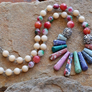 Può includere: Una collana fatta a mano con perline color crema, rosse e verdi, e dettagli argentati. La collana ha un design unico con pendenti in pietra allungati e colorati. La collana è esposta su una superficie di pietra.