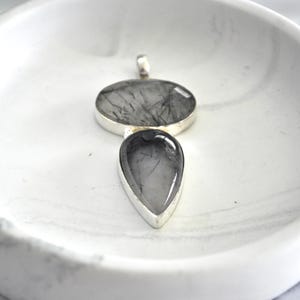 May include: A silver pendant featuring two black and clear quartz stones. The top stone is oval shaped and the bottom stone is teardrop shaped.