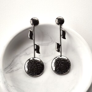 Puede incluir: Un par de pendientes negros colgantes con un disco redondo texturizado y tres abalorios en forma de hoja que cuelgan de cada pendiente.