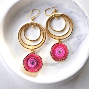 Puede incluir: Pendientes de aro dorados con un colgante de piedra de ágata rosa. Los pendientes tienen tres círculos concéntricos.