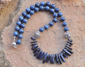Puede incluir: Un collar de piedras preciosas azules con cuentas de plata y un cierre de plata. El collar presenta una hebra larga de cuentas redondas y una hebra más corta de cuentas puntiagudas.