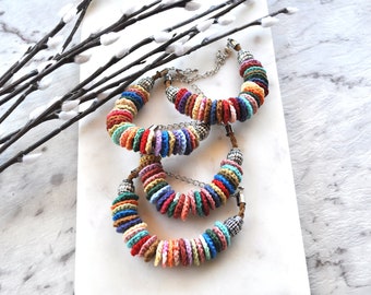 May include: Three colorful crochet bracelets with a variety of colors, including red, blue, orange, and green. The bracelets are arranged on a white marble surface. Each bracelet has a silver chain and decorative end caps.