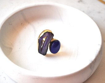 May include: A gold ring with a purple agate stone and a blue stone. The ring is on a white marble surface.