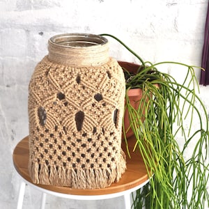 Könnte beinhalten: Eine Glasvase mit einer Makramee-Hülle, die ein geometrisches Muster und Fransen aufweist. Die Vase steht auf einem Holztisch mit einer Pflanze im Hintergrund.