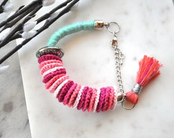 Könnte beinhalten: Ein gehäkeltes Armband in Rosa und Weiß. Das Armband hat einen silbernen Verschluss und einen rosafarbenen Quasten.