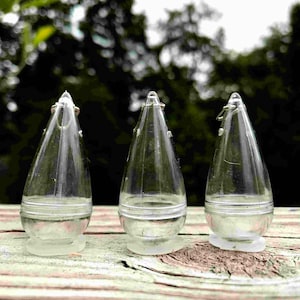 Puede incluir: Tres contenedores de plástico transparente en forma de lágrima con un pequeño lazo de metal en la parte superior. Los contenedores están sobre una superficie de madera.