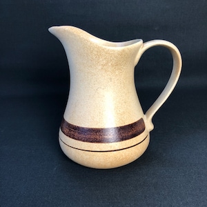May include: A tan ceramic pitcher with a curved handle and spout. The pitcher has a dark brown band around the middle and a speckled finish. The pitcher is likely used for serving beverages.