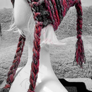 May include: A hand-crocheted hat in red, grey, and black, shown on a white mannequin head. The hat has a textured pattern and long, plaited tassels. This winter accessory is suitable for cold weather.