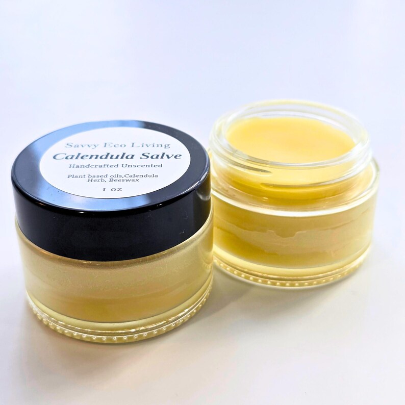 May include: A glass jar of yellow calendula salve with a black lid featuring a white label that reads Savvy Eco Living, Calendula Salve, Handcrafted Unscented, Plant based oils, Calendula Herb, Beeswax, 1 OZ. An open jar sits beside it.