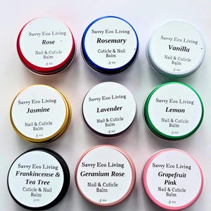 May include: Nine small, round metal tins of nail and cuticle balm are arranged on a white surface. Each tin has a different colored lid and a label with the scent name, such as Rose, Rosemary, and Vanilla. Each tin contains .5 oz of balm.