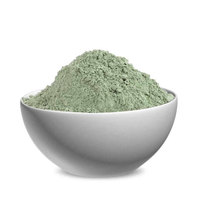May include: A smooth white ceramic bowl filled with a mound of fine, light sage green powder. The powder has a dry, textured appearance with small clumps, piled high above the rim of the bowl, creating a distinct contrast against the white container.