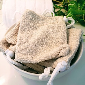 May include: A collection of beige textured bath mitts with white rope handles and white plastic toggles, arranged in a white bowl. A white washcloth with a rope handle is in the background. Green leaves are visible in the background.