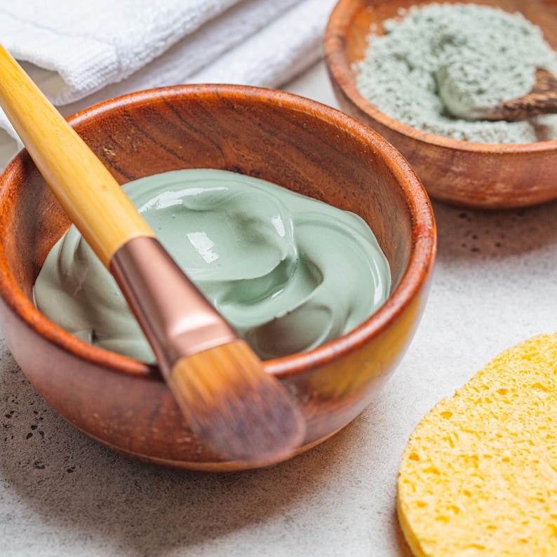 May include: A wooden bowl containing a smooth, light green facial mask with a wooden-handled applicator brush resting inside. A second wooden bowl holds dry green clay powder, and a round yellow sponge sits nearby for skincare application.