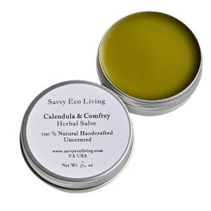 May include: A silver tin with a white label that reads "Savvy Eco Living Calendula & Comfrey Herbal Salve 100% Natural Handcrafted Unscented www.savvyecoliving.com VA USA Net Wt 2 oz". The tin contains a yellow salve.