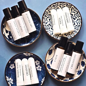 May include: An overhead shot features four small, decorative blue and white bowls, each holding aromatherapy products. The bowls are arranged on a blue surface. The products include essential oil roll-ons and aromatherapy inhalers, each labeled with different wellness-related names.