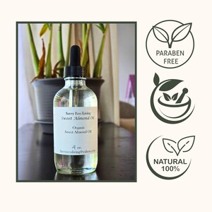 May include: A clear glass bottle with a dropper filled with organic sweet almond oil. The bottle has a white label with the text "Savvy Eco Living Sweet Almond Oil" and "Organic Sweet Almond Oil". The bottle is sitting on a wooden surface. The image also includes three green icons with the text "PARABEN FREE", "NATURAL 100%", and a mortar and pestle.
