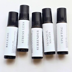 May include: Five essential oil roll-on bottles with black caps and white labels. The labels read "RELEASE", "STRESS LESS", "SURRENDER", "TRANQUIL NIGHTS", and "UPLIFTING". The bottles are arranged on a white surface.
