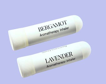 May include: Two white aromatherapy inhalers with rounded ends. One is labeled "BERGAMOT Aromatherapy Inhaler," and the other is labeled "LAVENDER Aromatherapy Inhaler." The background is a light purple color.