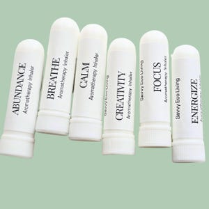 May include: Six white aromatherapy inhalers with black text. The inhalers are labeled with the words: ABUNDANCE, BREATHE, CALM, CREATIVITY, FOCUS, and ENERGIZE. The brand name "Savvy Eco Living" is also printed on some of the inhalers.