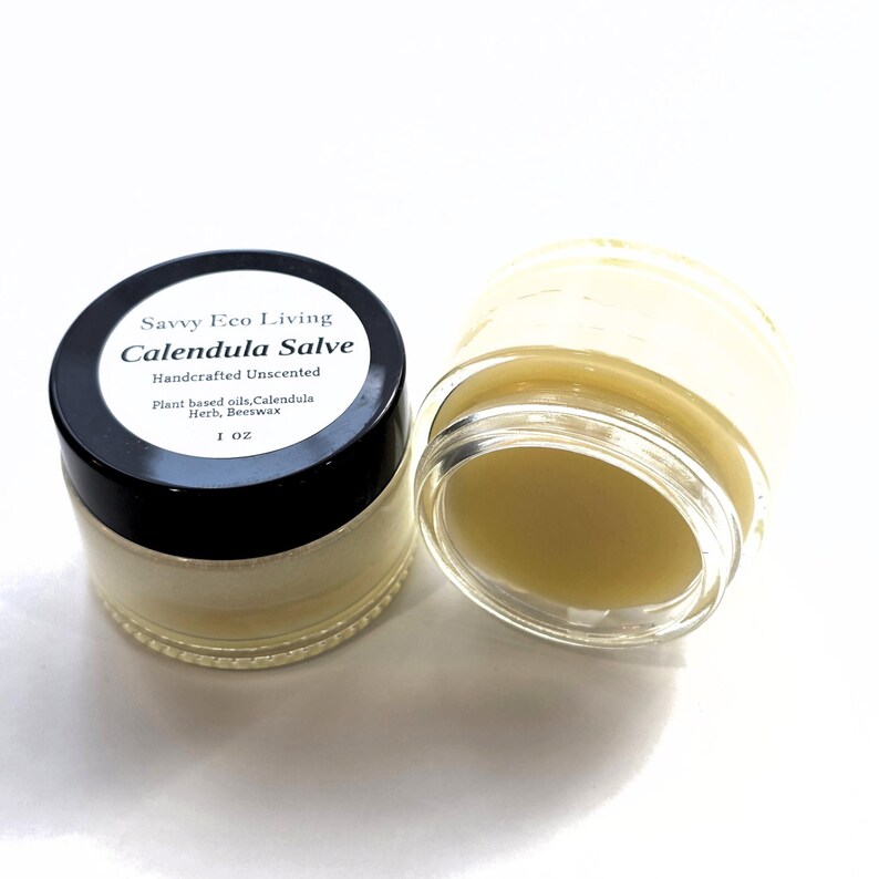 May include: Two small glass jars containing a pale yellow calendula salve. One jar has a black lid with a white label reading Savvy Eco Living, Calendula Salve, Handcrafted Unscented, Plant based oils, Calendula Herb, Beeswax, 1 OZ. The other jar is open.